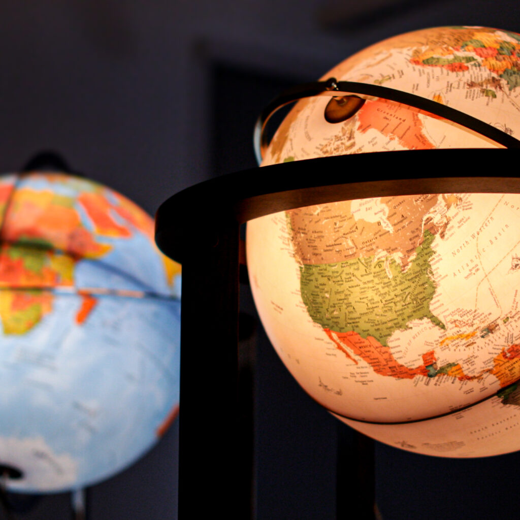 Zbliżenie na dwa podświetlane globusy; globus na pierwszym planie pokazuje Amerykę Północną, Amerykę Środkową i część Ameryki Południowej, z widocznymi nazwami krajów i granicami. Tło globusa jest rozmyte i przedstawia część Afryki i Europy. Otoczenie jest słabo oświetlone.