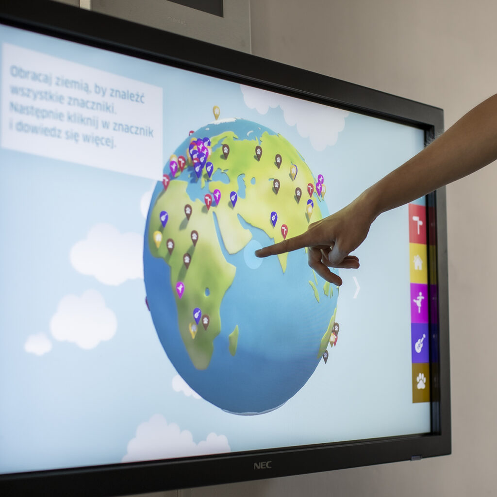Ręka wskazuje na cyfrową kulę ziemską na monitorze dotykowym, wyświetlając różne kolorowe znaczniki mapy na różnych kontynentach. Ekran pokazuje część polskiej instrukcji i ikony nawigacyjne po prawej stronie. Tło jest jasne z chmurami.