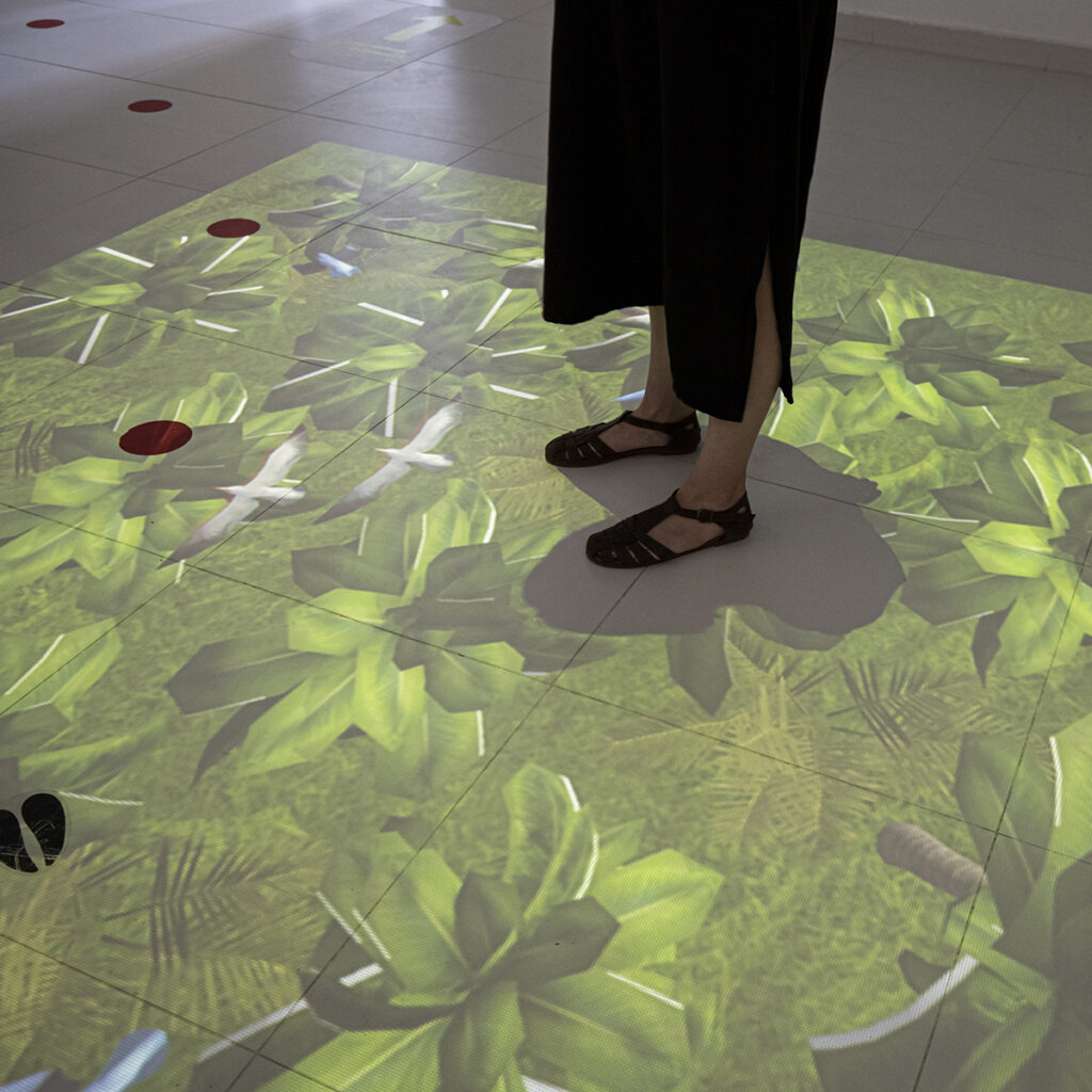 Osoba w czarnej spódnicy i sandałach stoi na wyłożonej kafelkami podłodze z jasną cyfrową projekcją zielonych liści, ptaków i roślin. Czerwone kropki i małe czarne ślady stóp są również widoczne na błyszczących białych kafelkach wokół projekcji.