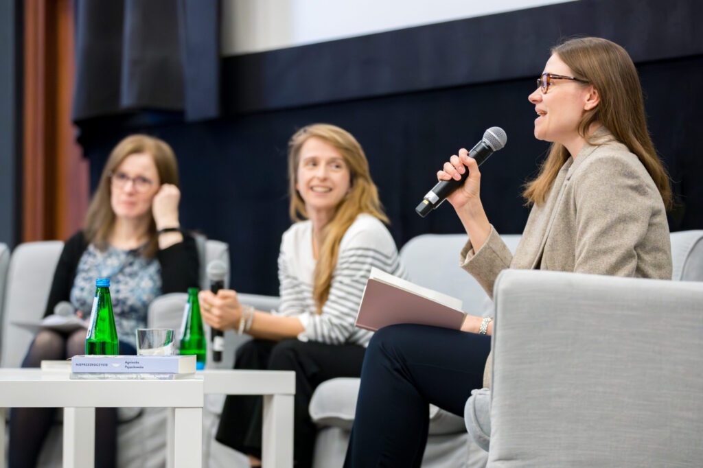 Trzy kobiety siedzą na scenie w fotelach podczas dyskusji panelowej. Jedna z nich, trzymając mikrofon i dokumenty, przemawia. Pozostałe dwie uważnie słuchają. Na pierwszym planie znajduje się stół z butelkami wody i szklankami. Wszystkie trzy wyglądają na zaangażowane i profesjonalne.