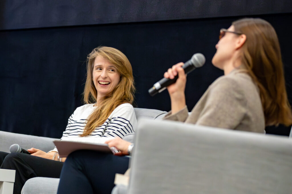 Dwie kobiety siedzą na jasnoszarych krzesłach i prowadzą rozmowę. Jedna z nich, ubrana w koszulę w paski, uśmiecha się ciepło, podczas gdy druga, w beżowej kurtce i okularach przeciwsłonecznych, mówi do mikrofonu. Tło jest ciemne i proste.