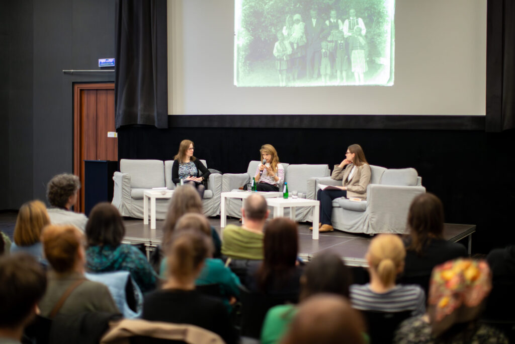 Trzy kobiety siedzą na scenie w fotelach, przemawiając do publiczności. Za nimi na ekranie wyświetlane jest czarno-białe zdjęcie rodziny. Publiczność zwrócona jest w stronę sceny, słuchając uważnie w nowoczesnej sali konferencyjnej z ciemnymi ścianami i zamkniętymi drzwiami.