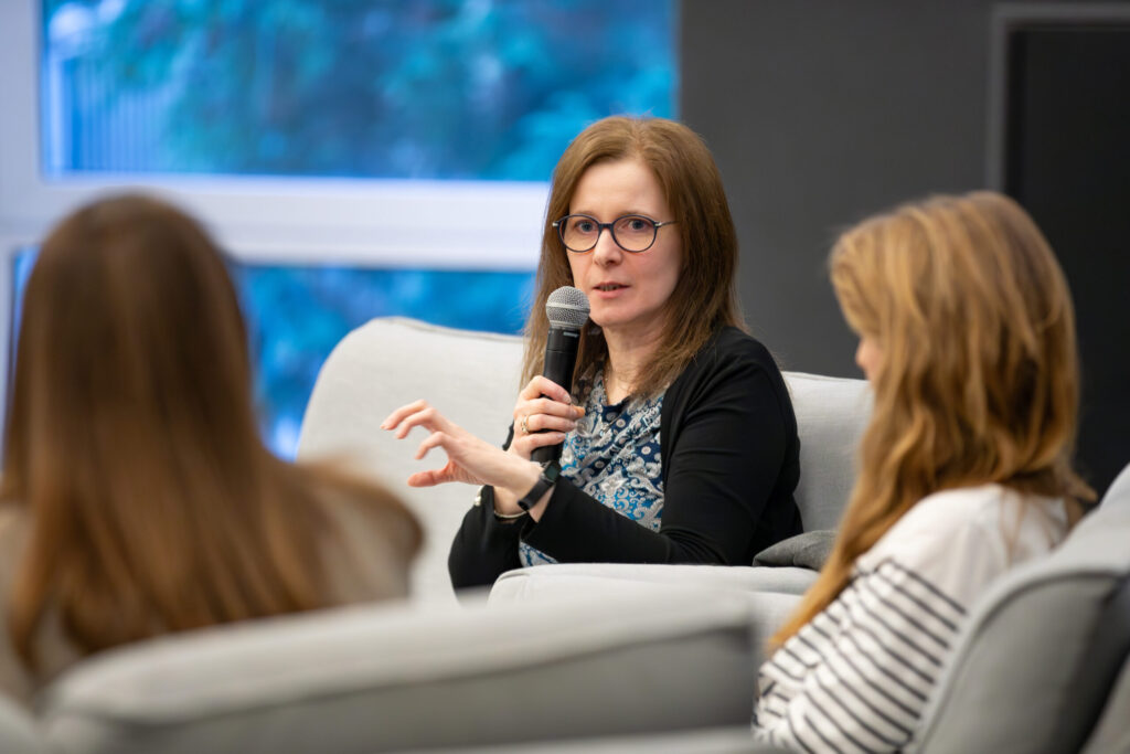 Kobieta w okularach mówi do mikrofonu, gestykulując ręką. Siedzi na jasnej sofie między dwiema innymi kobietami, które są zwrócone w jej stronę. Tło jest rozmyte, ukazując okno i zieleń na zewnątrz.