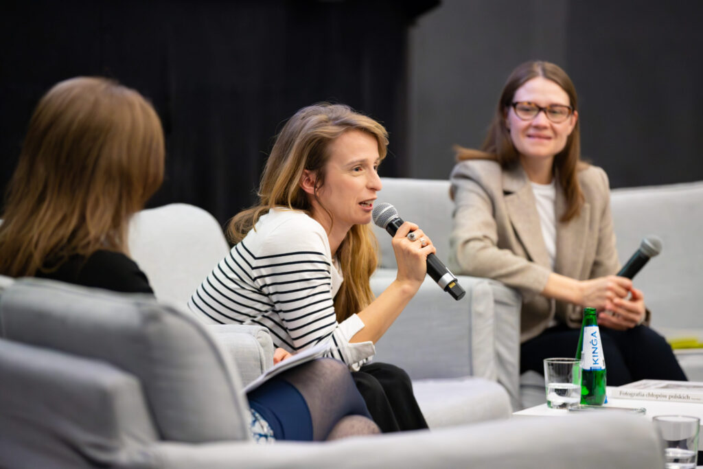 Trzy kobiety siedzą na jasnoszarych krzesłach w panelu dyskusyjnym. Kobieta w centrum, trzymająca mikrofon, mówi, podczas gdy pozostałe słuchają. Jedna z kobiet ma notatnik, a druga nosi okulary i beżową marynarkę. Na stole przed nimi stoi zielona butelka.
