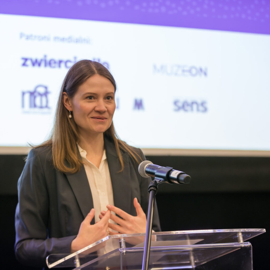 Kobieta o prostych brązowych włosach, ubrana w czarną marynarkę i białą koszulę, przemawia na podium z mikrofonem. Za nią znajduje się ekran wyświetlający kilka logo i tekst w języku polskim. Uśmiecha się i gestykuluje rękami.