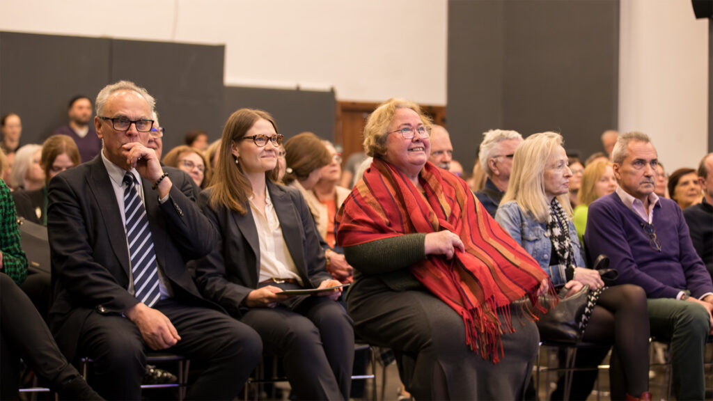 Zróżnicowana grupa dorosłych siedzi w rzędach, uważnie obserwując coś poza kamerą. W pierwszym rzędzie mężczyzna w garniturze, dwie kobiety - jedna uśmiechnięta z czerwonym szalikiem - oraz inne osoby słuchają i angażują się, sugerując formalne wydarzenie lub wykład w dobrze oświetlonym pomieszczeniu.