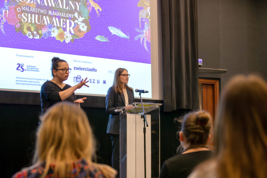 Kobieta w garniturze przemawia na podium w sali konferencyjnej. Obok niej gestykuluje tłumacz języka migowego. Kilka osób siedzi na widowni. Na ekranie za nimi wyświetlany jest kolorowy slajd z wydarzenia.