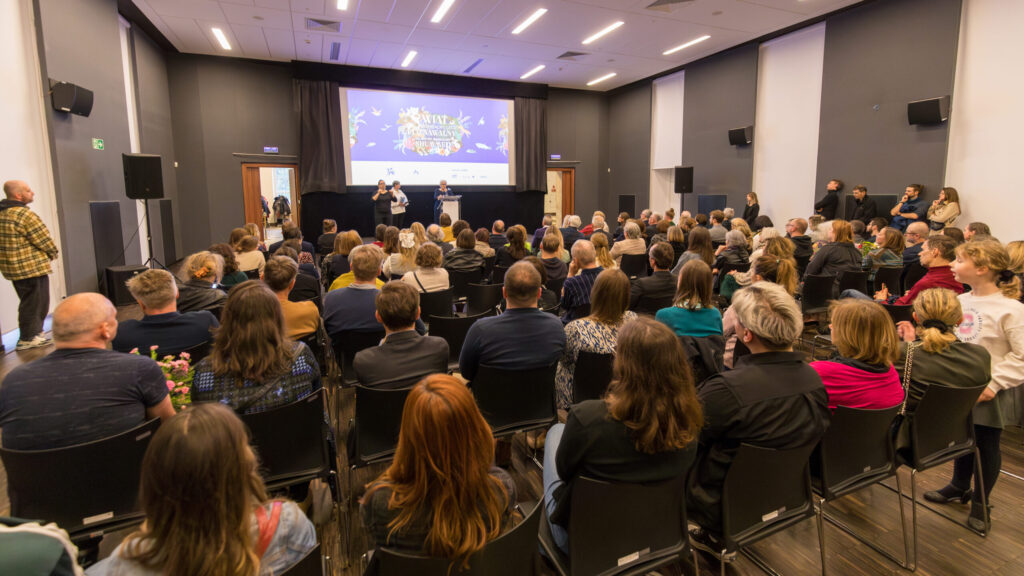 Duża publiczność siedzi w rzędach naprzeciwko sceny, na której dwóch prelegentów stoi przed ekranem wyświetlającym "Nature Festival". Pomieszczenie jest dobrze oświetlone, z ciemnymi ścianami, a kilka osób stoi po bokach i z tyłu, obserwując prezentację.