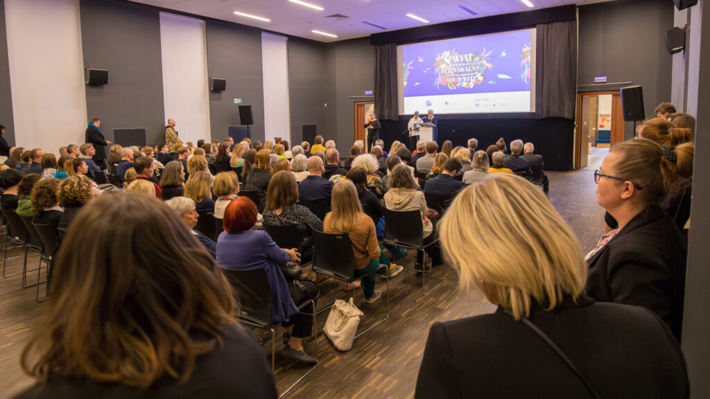 Duża publiczność siedzi w rzędach naprzeciwko sceny w sali konferencyjnej. Na scenie prelegenci stoją na podium obok ekranu wyświetlającego kolorowe grafiki i tekst. Pomieszczenie jest dobrze oświetlone, a uczestnicy uważnie obserwują prezentację.