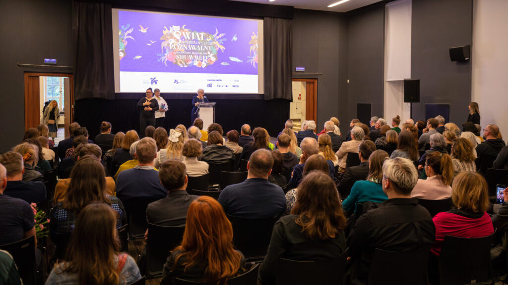 Duża publiczność siedzi naprzeciwko sceny, na której dwie osoby stoją na podium i przemawiają. Za nimi znajduje się kolorowy ekran wydarzenia z tekstem i logo. Pokój ma ciemne ściany, dwoje drzwi i jest jasno oświetlony, a ludzie uważnie słuchają mówców.