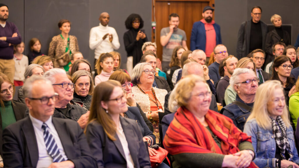 Zróżnicowana grupa dorosłych i kilkoro dzieci siedzi blisko siebie w rzędach, uważnie obserwując wydarzenie w dobrze oświetlonym pomieszczeniu. Niektóre osoby stoją z tyłu. Większość z nich nosi okulary i eleganckie ubrania, a ich miny wahają się od skupionych do uśmiechniętych.