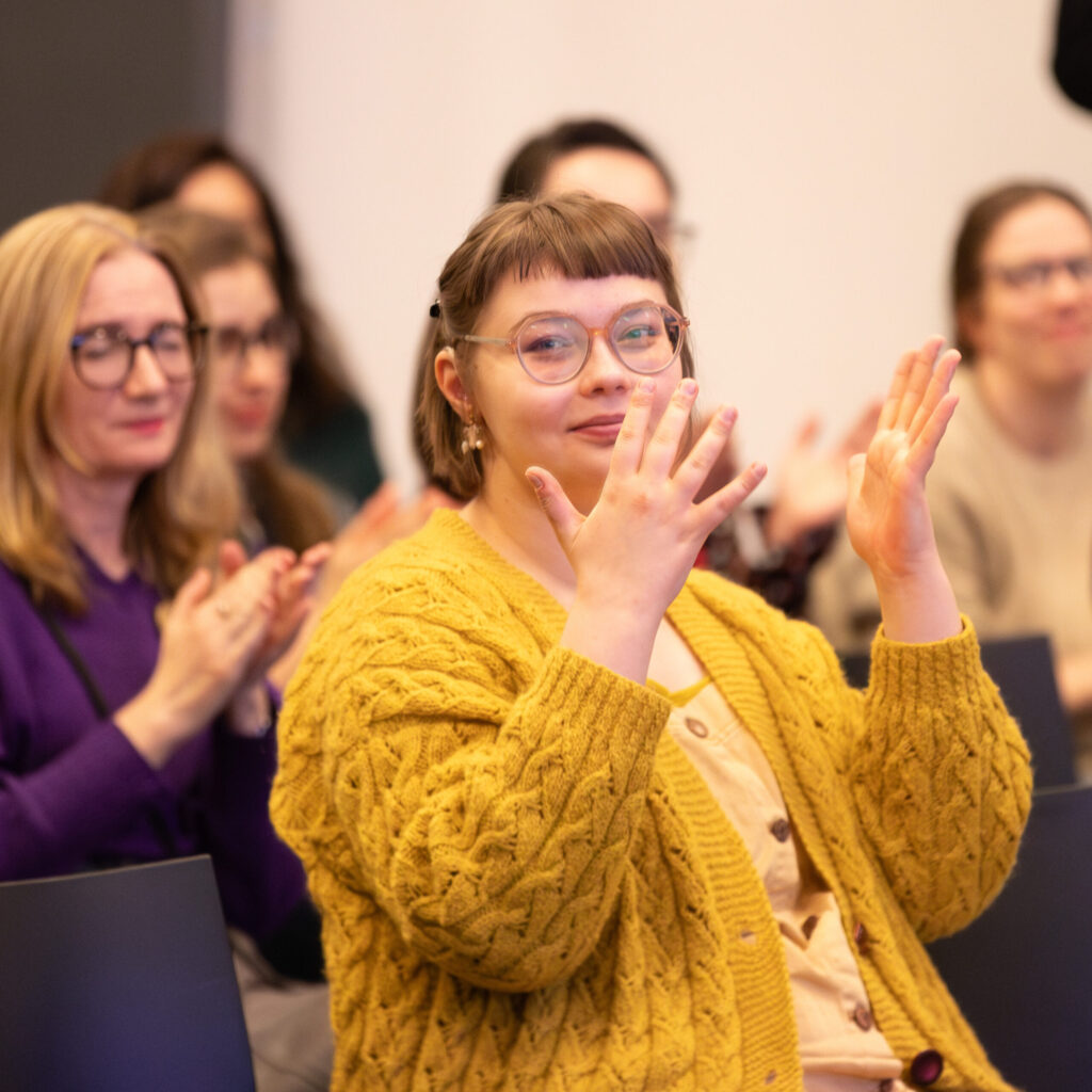 Młoda osoba z krótkimi brązowymi włosami i okularami, ubrana w żółty sweter z dzianiny, siedzi w grupie i gestykuluje podniesionymi rękami. Kilka osób w tle, głównie kobiet, klaszcze i uważnie obserwuje w dobrze oświetlonym pomieszczeniu.