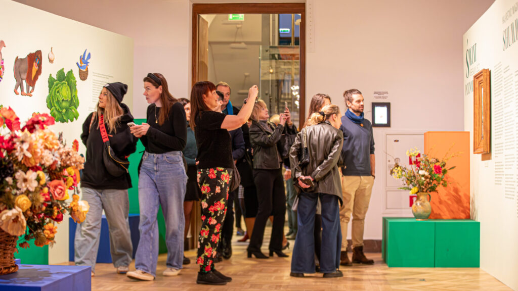 Grupa ludzi w swobodnych strojach obserwuje dzieła sztuki i teksty wyświetlane na jasno oświetlonych ścianach muzeum. Niektórzy robią zdjęcia, inni rozmawiają. W pomieszczeniu stoją wazony z kolorowymi kwiatami i zielone stojaki. Drzwi otwierają się na korytarz w tle.