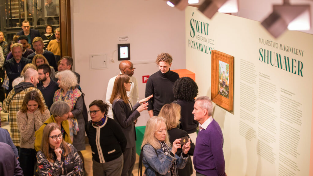 Ruchliwa galeria sztuki z różnymi ludźmi rozmawiającymi i oglądającymi wystawę zatytułowaną "Shimmer". Jasno oświetlona ściana wyświetla tekst i oprawiony obraz. Odwiedzający stoją, rozmawiają i robią zdjęcia. Atmosfera jest żywa i towarzyska.