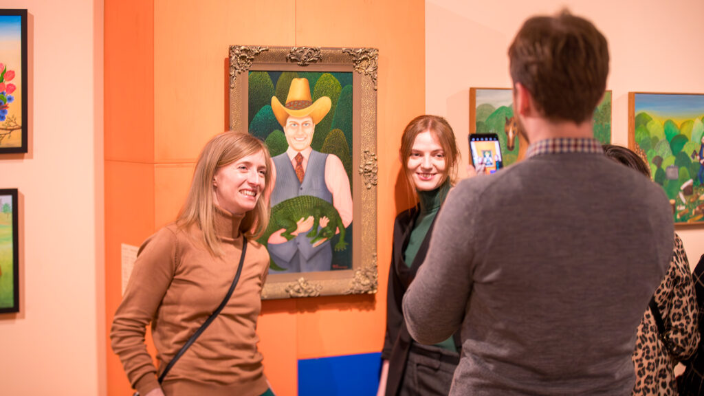 Dwie kobiety uśmiechają się i pozują do zdjęcia przed kolorowym obrazem przedstawiającym uśmiechniętego mężczyznę w żółtym kapeluszu i kamizelce trzymającego zielone zwierzę. Trzecia osoba, odwrócona tyłem do kamery, robi sobie zdjęcie smartfonem w otoczeniu galerii sztuki.