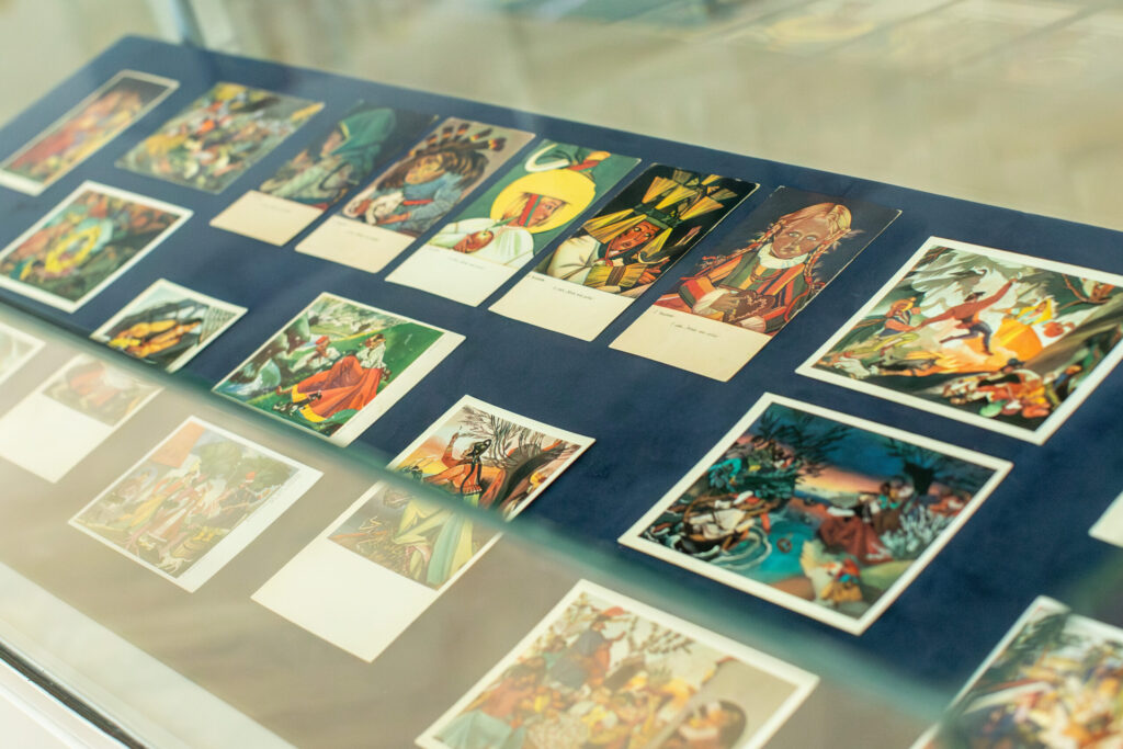 W gablocie znajdują się ilustrowane pocztówki i wydruki w stylu vintage, ułożone starannie na niebieskim materiale. Kolorowe obrazy przedstawiają różne osoby, sceny i czynności, a niektóre z nich zawierają odręczne podpisy lub notatki pod ilustracjami.