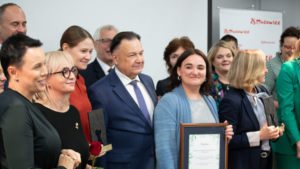 Grupa ludzi, ubranych w formalne stroje, stoi razem na imprezie wewnętrznej. Niektórzy trzymają oprawione certyfikaty i czerwoną różę. Grupa wydaje się świętować osiągnięcie. W tle widoczny jest biały baner z czerwonym logo "Mazowsza".