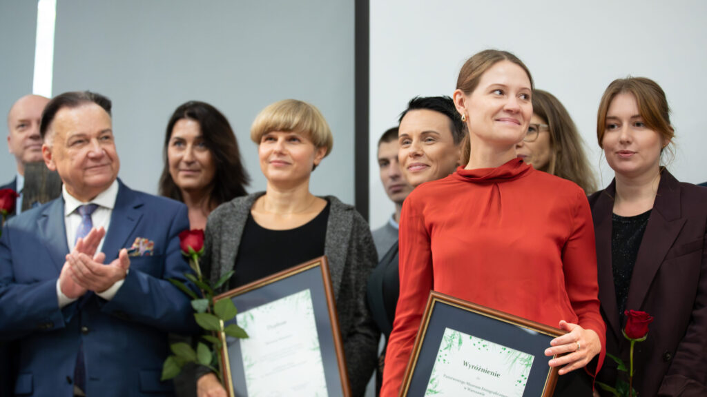 Grupa ludzi na ceremonii wręczenia nagród. Dwie kobiety z przodu trzymają oprawione certyfikaty i róże, uśmiechając się. Jedna ma na sobie czerwoną bluzkę; druga ma krótkie blond włosy i czarny strój. Inni w formalnych strojach stoją z tyłu, niektórzy się uśmiechają, jeden mężczyzna klaszcze.