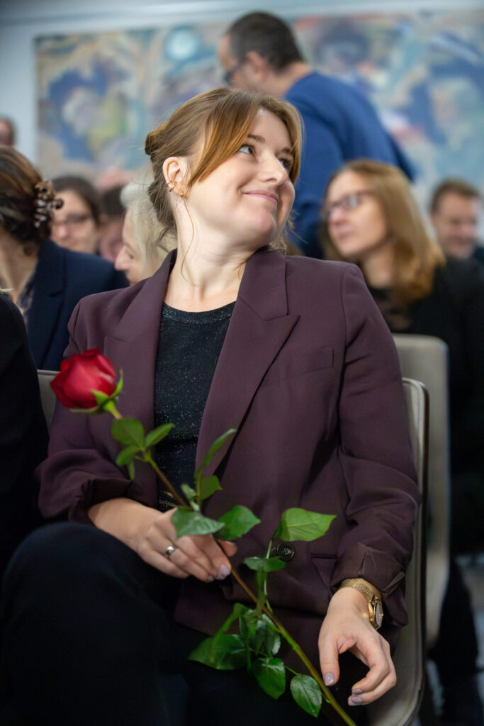 Kobieta z jasnobrązowymi włosami w luźnym upięciu, ubrana w bordową marynarkę i czarną koszulę, uśmiecha się, trzymając czerwoną różę. Siedzi wśród grupy ludzi, wielu niewyraźnych w tle, co wydaje się być formalnym lub uroczystym wydarzeniem.