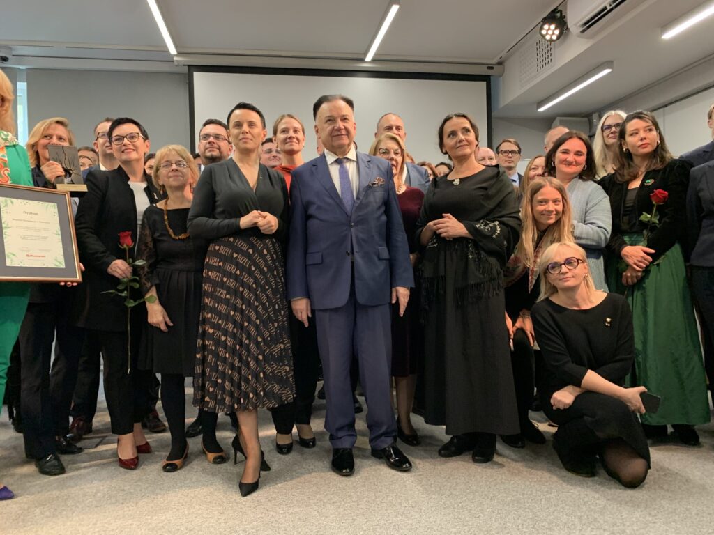 Duża grupa ludzi, ubranych formalnie, stoi razem w pomieszczeniu do zdjęcia. Niektórzy trzymają czerwone róże i oprawiony certyfikat. Centralny mężczyzna ma na sobie niebieski garnitur i krawat. Grupa wygląda na szczęśliwą, pozując na tle białych ścian i ekranu projekcyjnego.