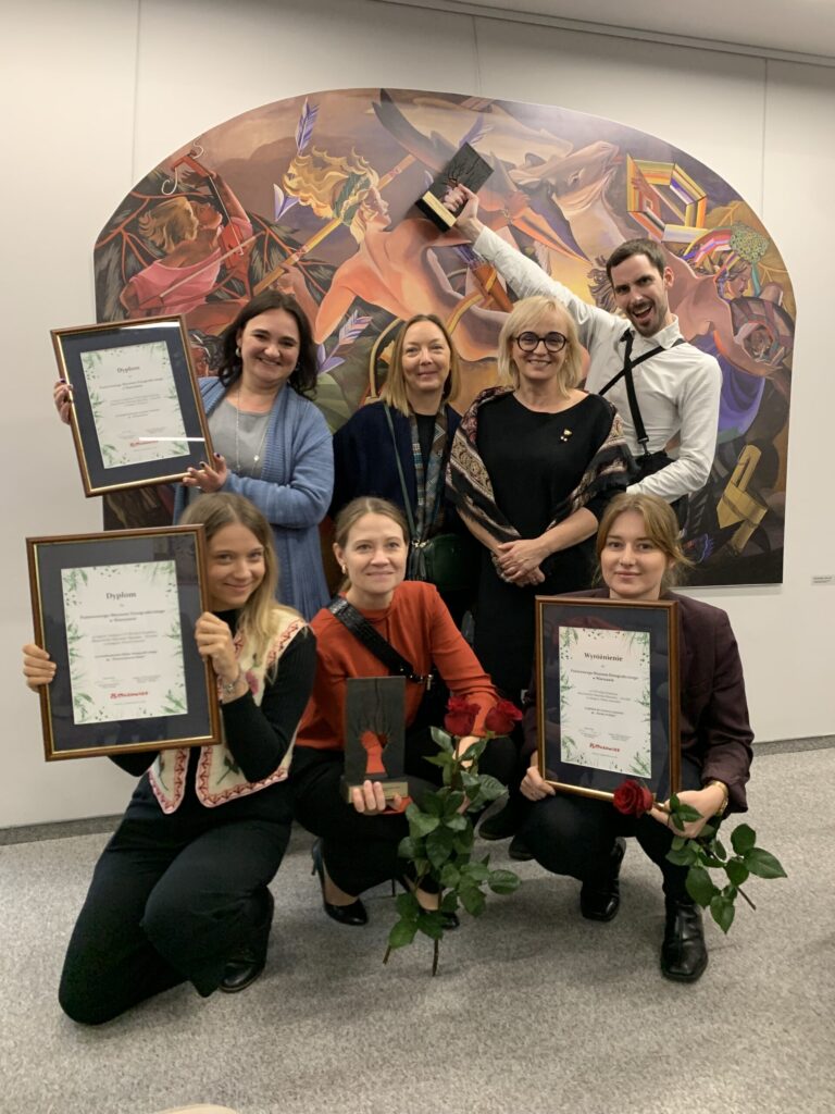 Siedem osób pozuje w pomieszczeniu przed kolorowym abstrakcyjnym muralem. Cztery klęczą trzymając certyfikaty i róże, podczas gdy trzy stoją za nimi uśmiechając się, a jedna podnosi certyfikat w geście świętowania. Wszyscy ubrani są w półformalne stroje, co sugeruje, że jest to wydarzenie związane z nagrodą lub uznaniem.