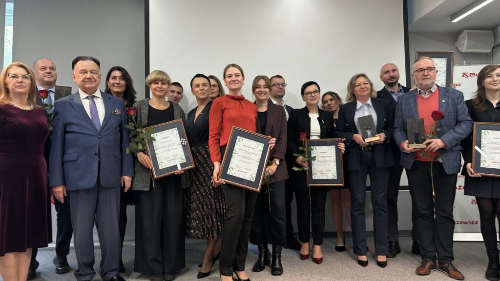 Grupa trzynastu dorosłych, ubranych w formalne stroje, stoi pozując w pomieszczeniu. Kilka osób trzyma certyfikaty i czerwone róże, a niektórzy trzymają trofea. Stoją przed pustym ekranem projekcyjnym, uśmiechając się, jakby świętowali osiągnięcie lub ceremonię wręczenia nagród.