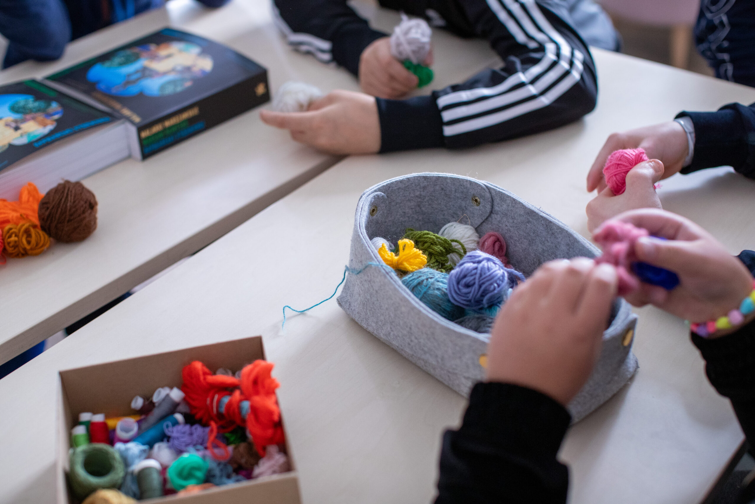 Kilka osób siedzi wokół stołu i zajmuje się kolorowymi kulkami z włóczki i pomponami. Na stole znajduje się szary filcowy pojemnik z różnymi włóczkami, a także książki i kartonowe pudełko z materiałami rzemieślniczymi. Widoczne są tylko ręce i ramiona uczestników.