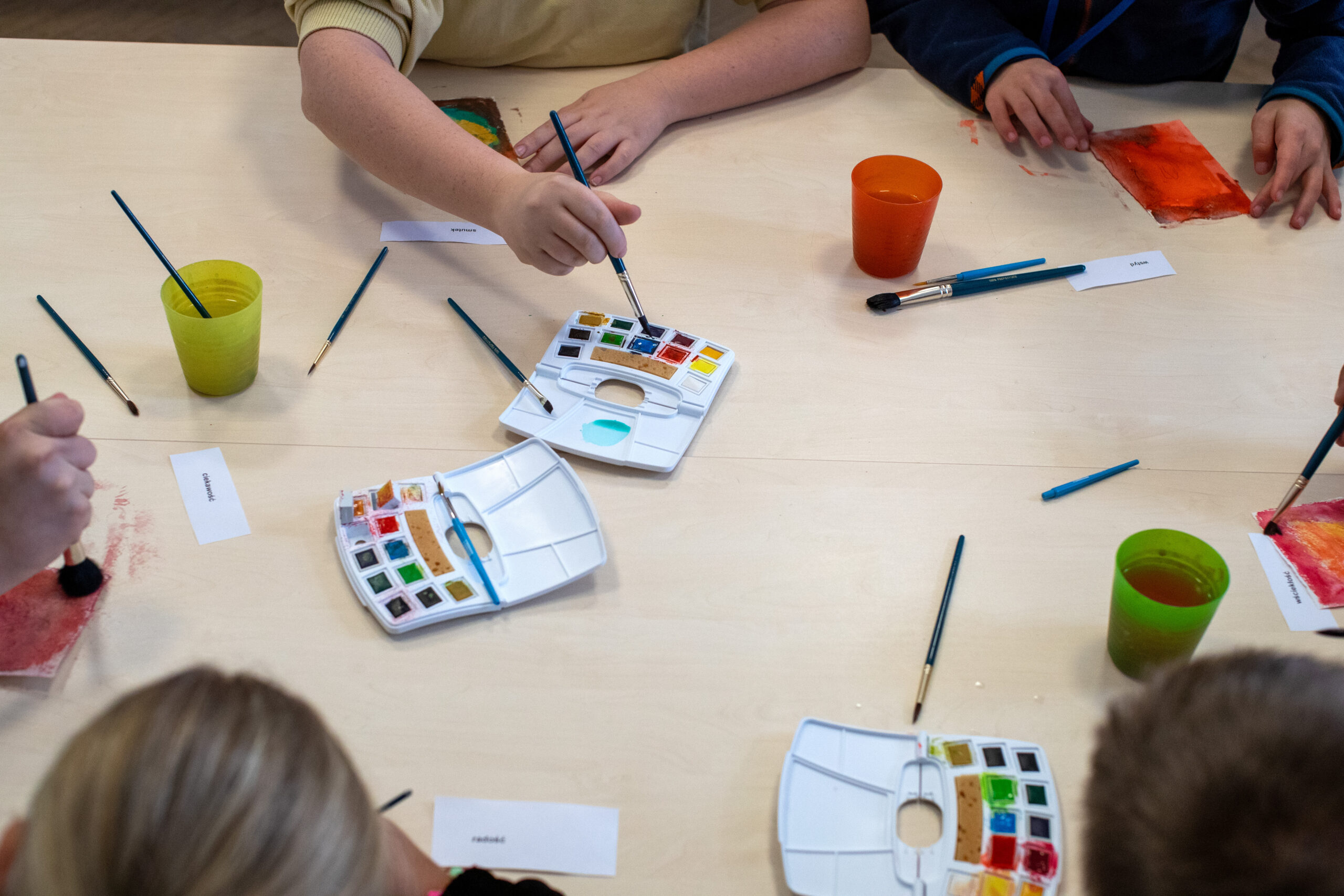 Kilka osób siedzi przy stole i maluje akwarelami. Widoczne są ich dłonie, pędzle i kubki z wodą. Każda osoba pracuje nad kolorowym obrazem, z odcieniami czerwieni i pomarańczy, podczas gdy kartki z imionami i nieużywane pędzle są rozrzucone na stole.