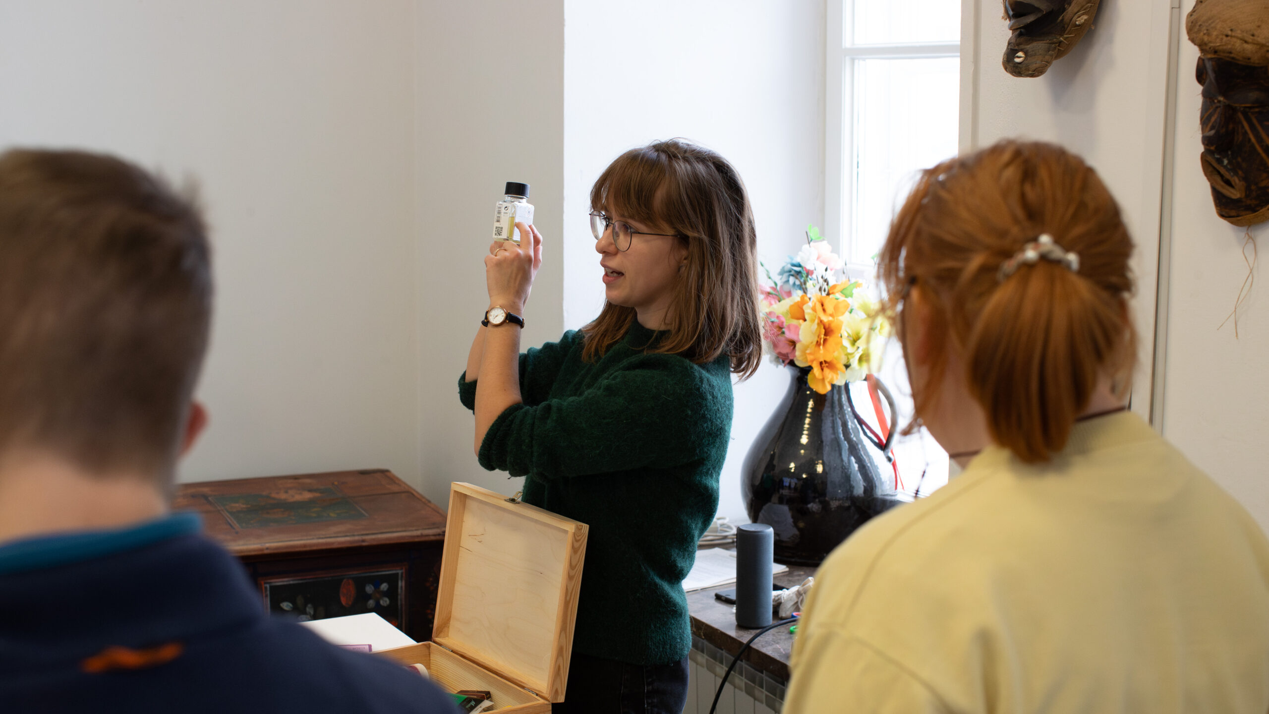 Kobieta w okularach i brązowych włosach trzyma małą butelkę, rozmawiając z dwiema osobami odwróconymi plecami do kamery. Stoi przy stole z drewnianą skrzynką, w pokoju z wazonem kolorowych kwiatów w pobliżu jasnego okna.