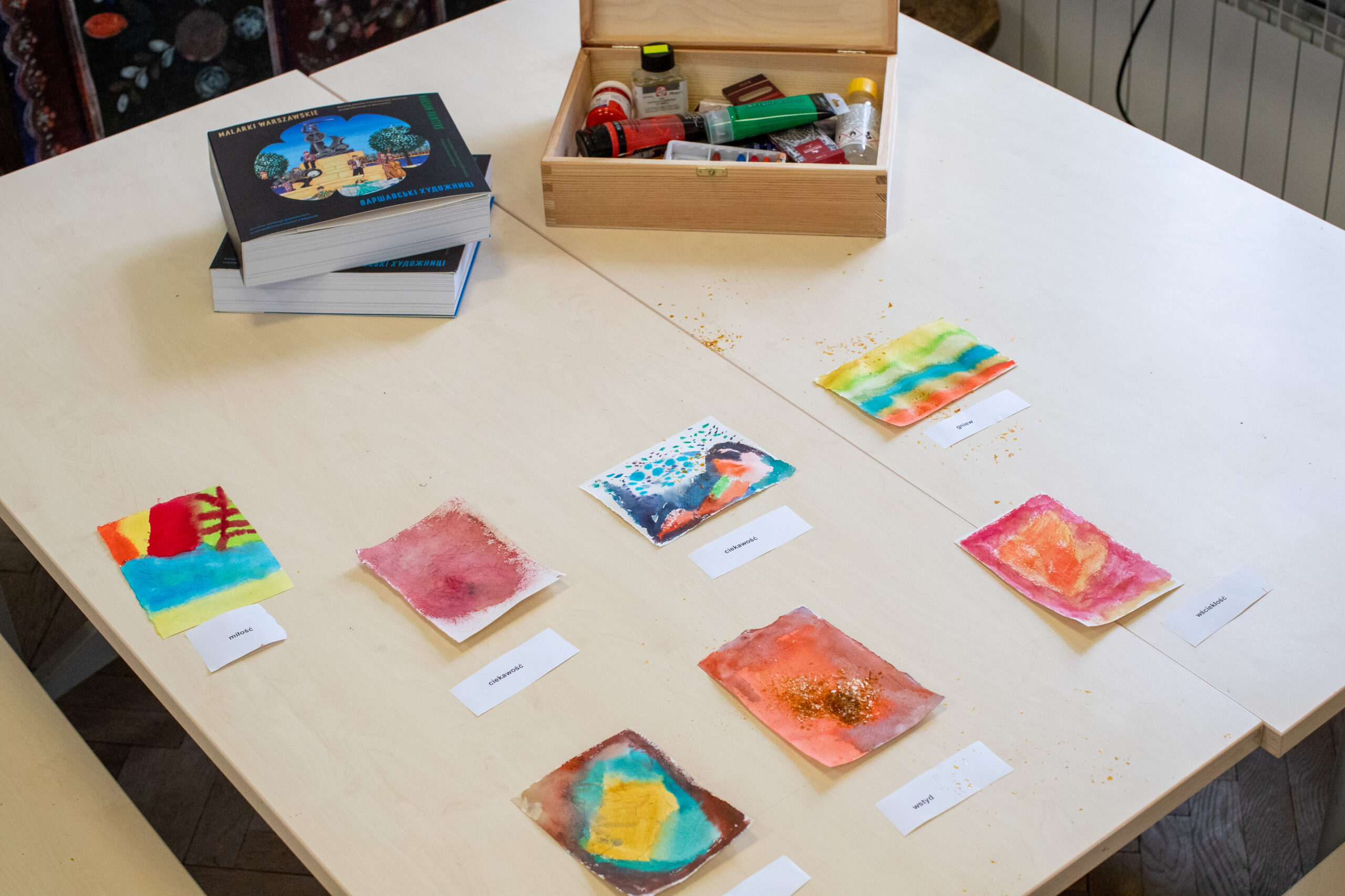 Na jasnym drewnianym stole znajduje się sześć małych, kolorowych akwareli na papierze, każdy oznaczony białą kartką. Z tyłu stoi otwarta drewniana skrzynia wypełniona przyborami plastycznymi i stosem dużych ilustrowanych książek. Scena wygląda na kreatywną i zorganizowaną.
