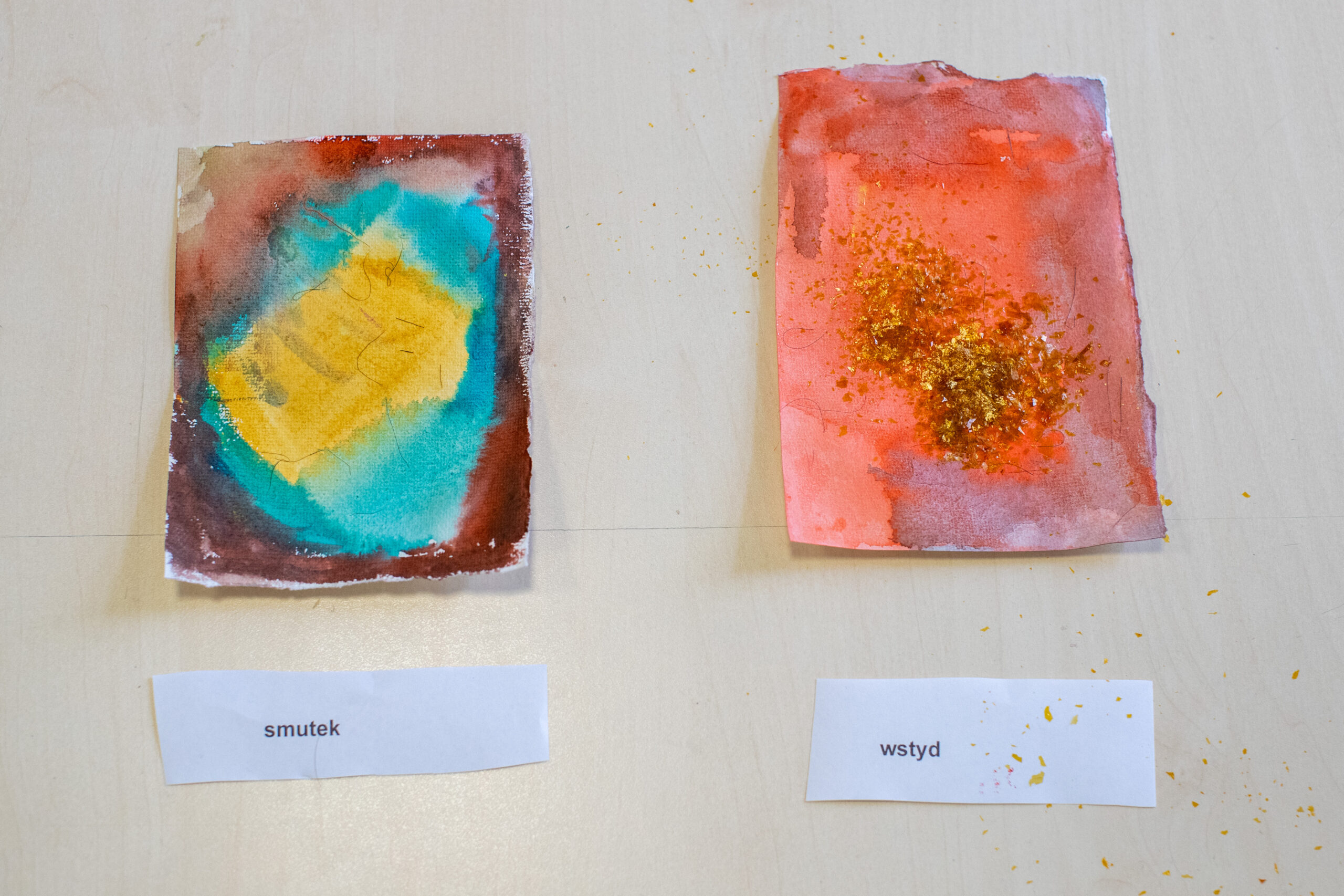 Dwa małe abstrakcyjne obrazy akwarelowe na jasnej powierzchni. Lewy obraz łączy kolory żółty, turkusowy i brązowy i jest oznaczony jako "smutek". Prawy obraz jest głównie czerwono-pomarańczowy ze złotym brokatem i jest oznaczony jako "wstyd". Obie etykiety są wydrukowane na białym papierze.