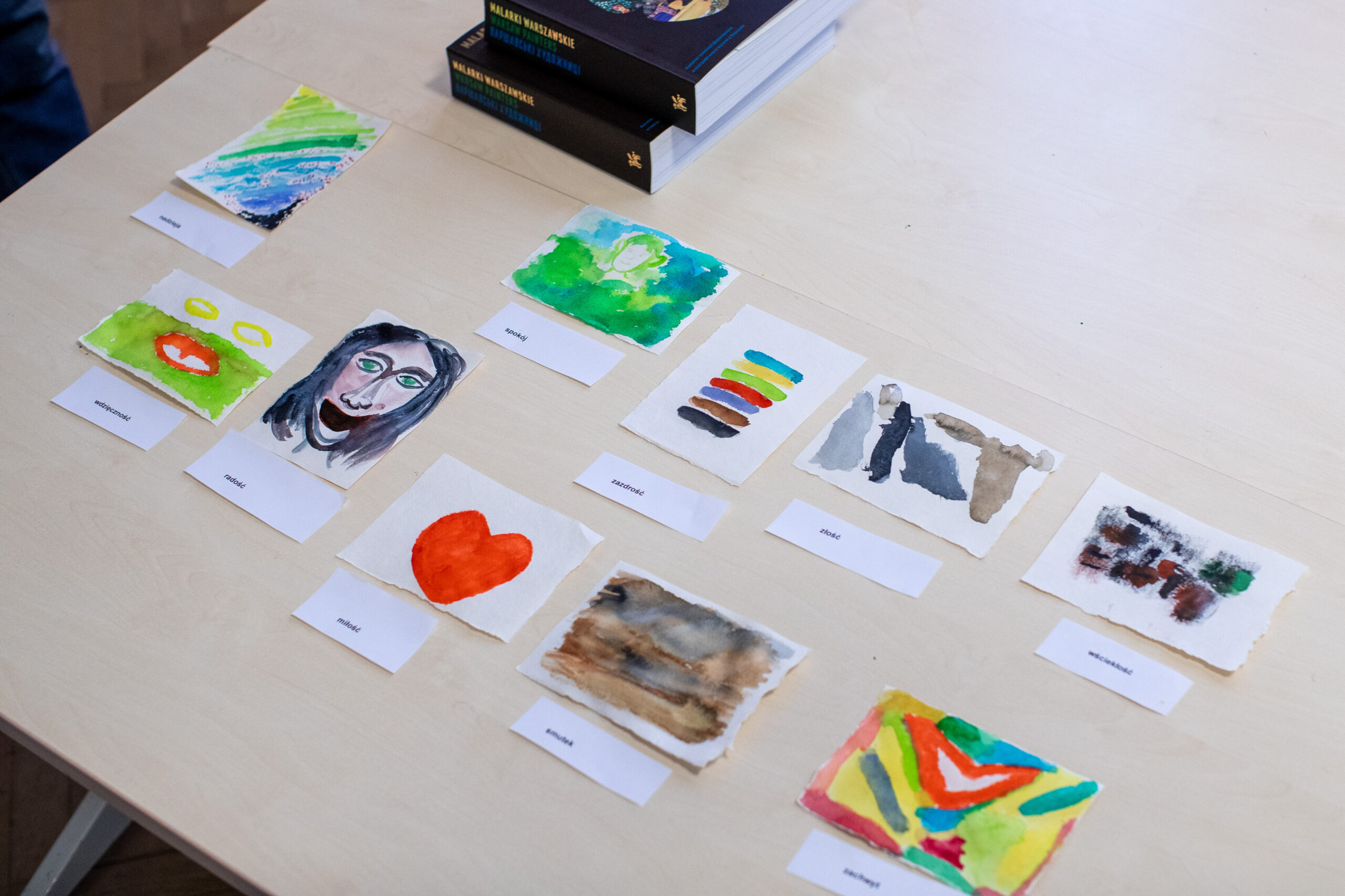 Na drewnianym stole znajduje się dziewięć małych abstrakcyjnych obrazów akwarelowych, każdy oznaczony karteczką. Nad dziełami sztuki znajdują się dwie ułożone w stos książki. Obrazy różnią się kolorem i stylem, w tym sercami, twarzami i abstrakcyjnymi kształtami.