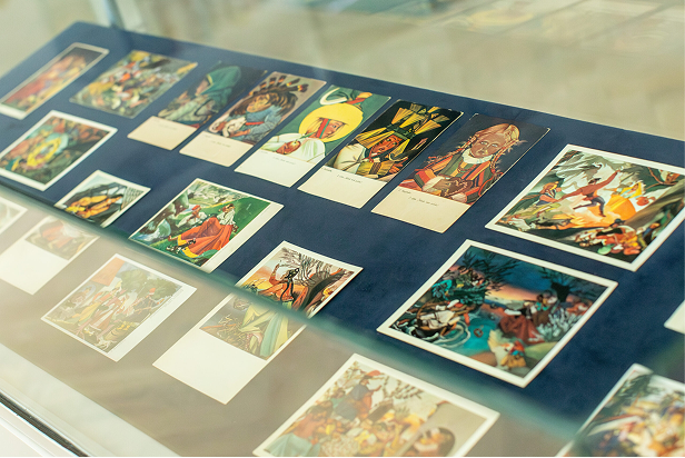 Szklana gablota przedstawia kolekcję kolorowych, ilustrowanych pocztówek w stylu vintage, ułożonych w rzędach na niebieskim materiale. Pocztówki przedstawiają różne sceny i postacie, w tym ludzi w kostiumach, żywe tła i żywą akcję.