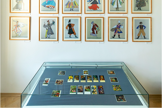 W szklanej gablocie na niebieskim materiale leżą kolorowe karty w stylu vintage. Za nią, na białej ścianie w trzech rzędach równomiernie rozmieszczono dwanaście oprawionych ilustracji przedstawiających ludzi w żywych, tradycyjnych lub teatralnych strojach.