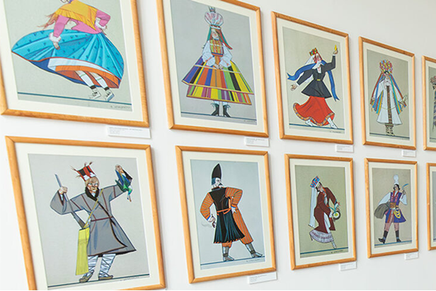 Ściana galerii przedstawia dwanaście oprawionych ilustracji ludzi w kolorowych, tradycyjnych strojach. Każda postać jest unikalnie ubrana, reprezentuje różne style i kultury, z dynamicznymi pozami. Ramki są ułożone w trzech rzędach na białej ścianie.