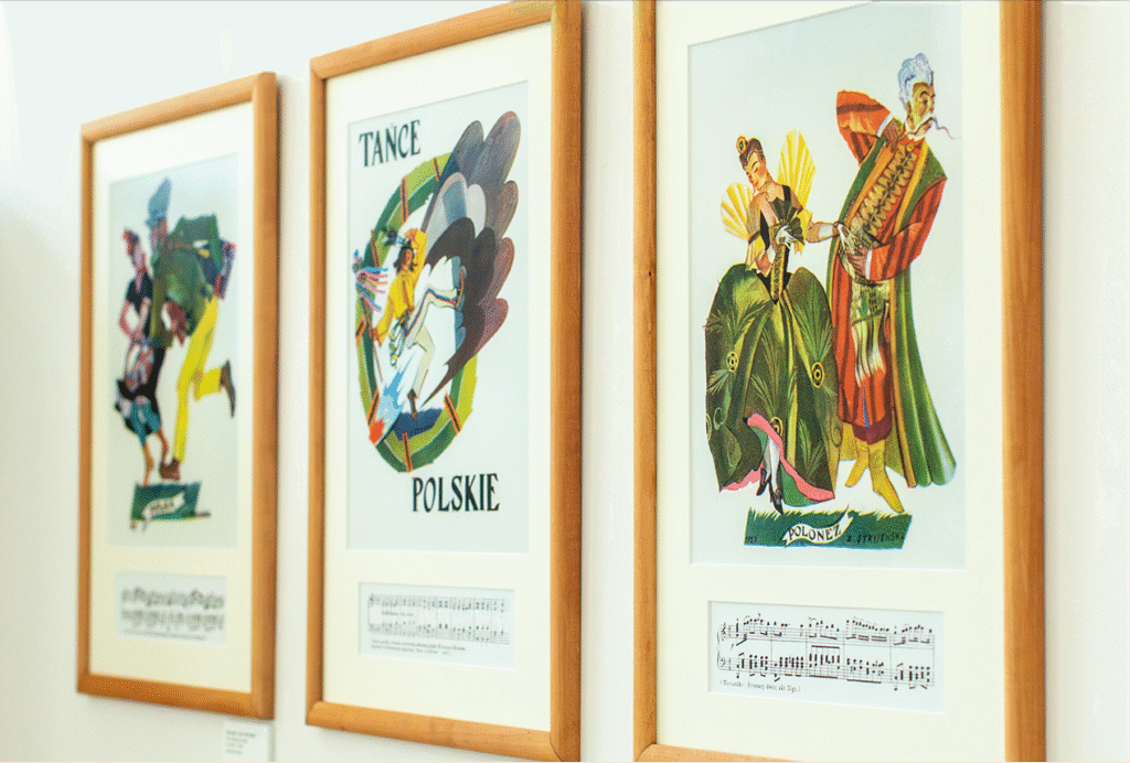 Na ścianie wiszą trzy oprawione plakaty, z których każdy przedstawia kolorowe ilustracje ludzi w tradycyjnych polskich strojach tanecznych. Na środkowym plakacie widnieje napis Tańce Polskie, a u dołu wszystkich umieszczono nuty. Ramy są wykonane z jasnego drewna.