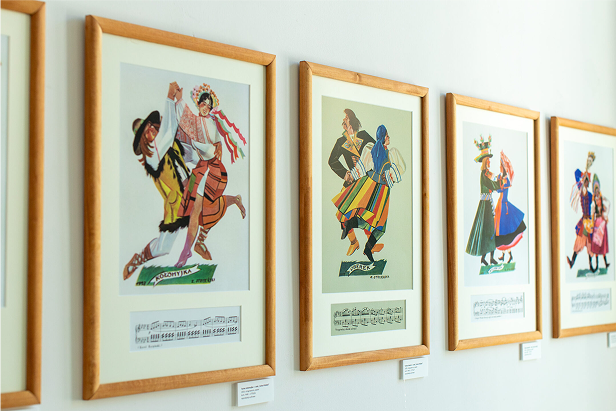 Na białej ścianie wyświetlane są oprawione ilustracje ludzi w kolorowych, tradycyjnych strojach ludowych. Każdy obraz zawiera muzykę pod tancerzami, a obrazy są oznaczone nazwami, sugerującymi różne tańce narodowe lub regionalne.