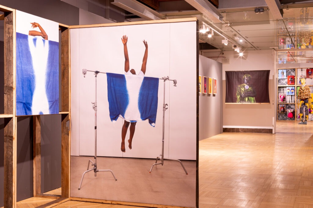 Galeria prezentuje sztukę nowoczesną; na pierwszym planie znajduje się duża fotografia osoby odwróconej tyłem, z podniesionymi rękami, udrapowana w niebieską tkaninę zawieszoną na stojakach. Inne dzieła sztuki i rzeźby są widoczne w tle pod oświetleniem szynowym na drewnianych podłogach.