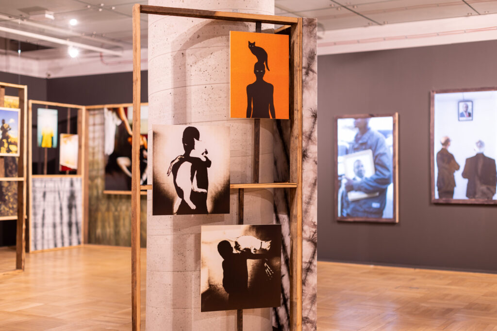 Drewniana konstrukcja ekspozycyjna stoi na filarze, trzymając trzy monochromatyczne dzieła sztuki przedstawiające sylwetki ludzi w różnych pozach. W tle widać więcej oprawionych zdjęć i dzieł sztuki w nowoczesnej galerii z drewnianymi podłogami i szarymi ścianami.