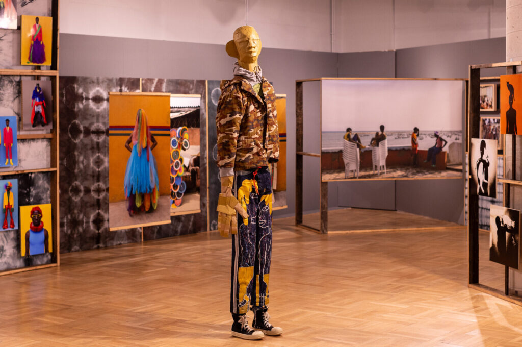 Manekin ubrany w kamuflażową kurtkę, kolorowe spodnie i czarne trampki stoi na wypolerowanej drewnianej podłodze w galerii sztuki. Na ścianach wiszą duże, żywe fotografie ludzi i abstrakcyjnych scen zamontowane w drewnianych ramach.