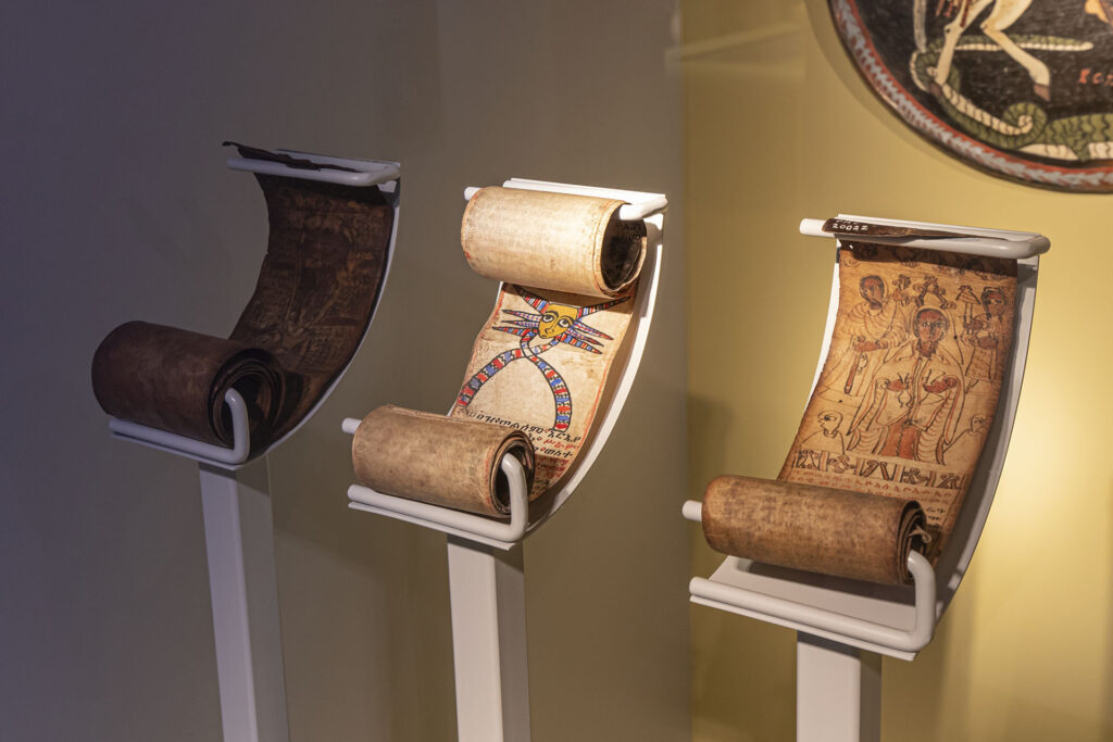 Trzy starożytne ilustrowane zwoje są wyświetlane pionowo na białych stojakach. Zwoje przedstawiają szczegółowe, kolorowe rysunki i tekst, przedstawiające różne sceny i postacie, z centralnym zwojem przedstawiającym skrzydlatą postać. Tło jest delikatnie oświetlone, podkreślając artefakty.