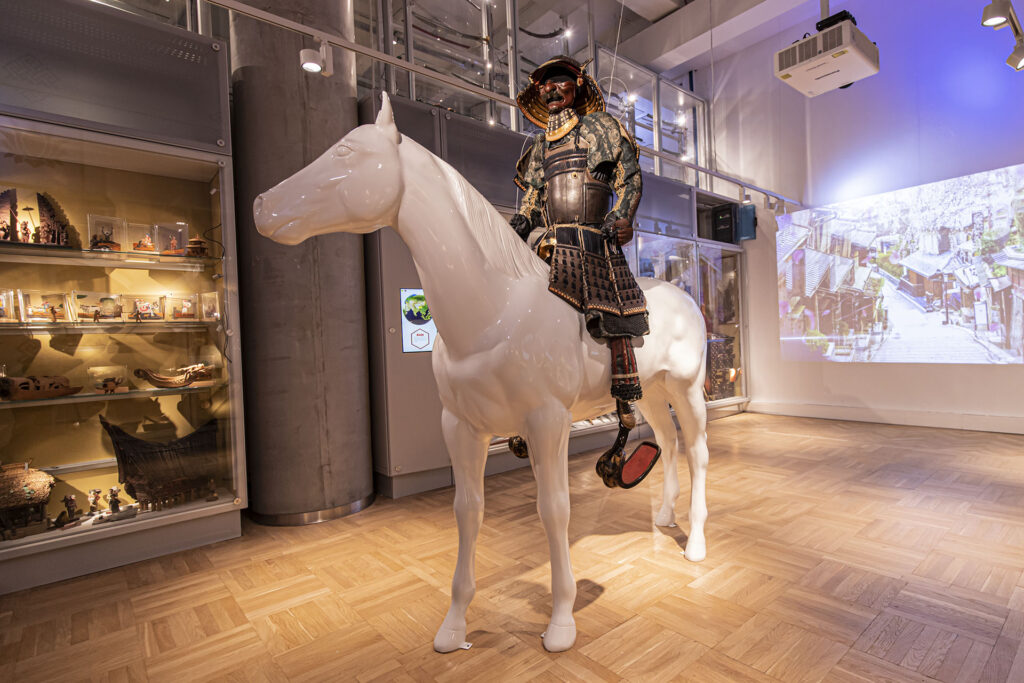 Eksponat muzealny przedstawia manekina w tradycyjnej japońskiej zbroi samurajskiej jadącego na dużym, białym, realistycznym posągu konia. Gabloty z artefaktami ustawione są wzdłuż ścian, a w tle widoczny jest duży wyświetlany obraz historycznej japońskiej sceny ulicznej.