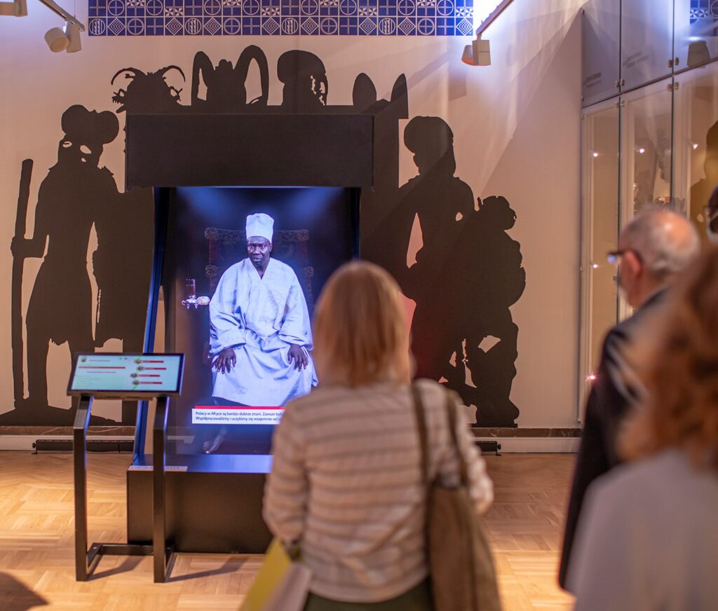 Trzy osoby stoją przed interaktywnym wyświetlaczem muzealnym przedstawiającym siedzącego mężczyznę w tradycyjnym białym stroju. Za wyświetlaczem znajdują się duże czarne sylwetki postaci w wyszukanych kostiumach, ustawione na tle oświetlonej galerii z drewnianymi podłogami i szklanymi gablotami.