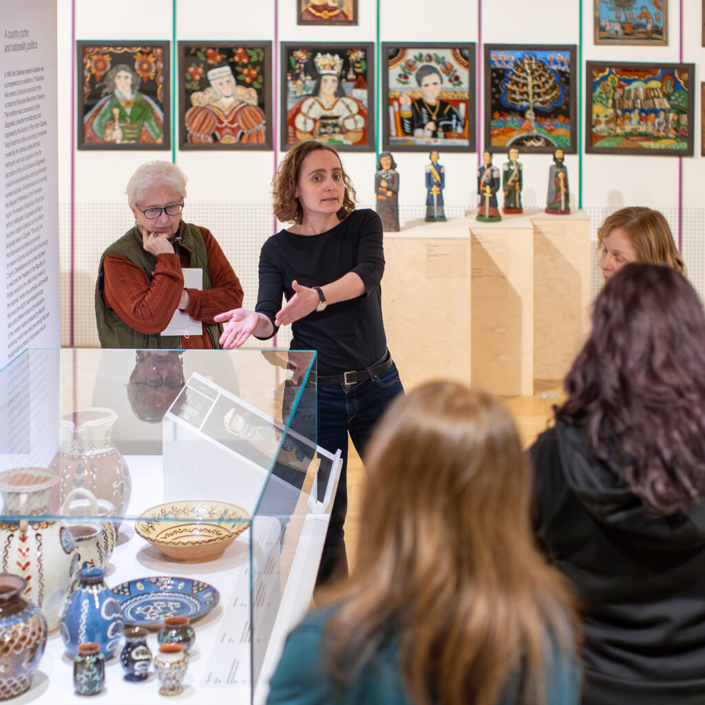 Kobieta gestykuluje, przemawiając do małej grupy w galerii muzeum. Stoją w pobliżu szklanej gabloty prezentującej zdobioną ceramikę, z kolorowymi ludowymi obrazami i figurkami na ścianach i postumentach za nimi. Inni uczestnicy uważnie słuchają.