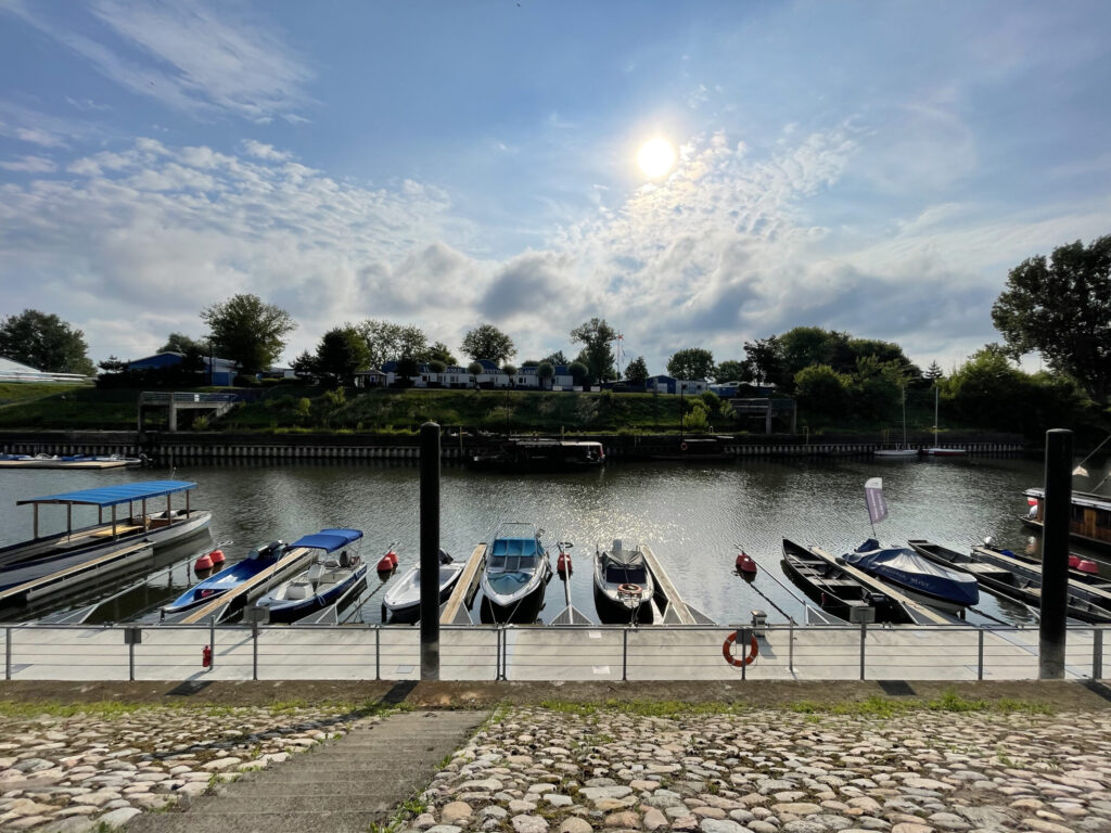 Mała przystań z kilkoma zadokowanymi łodziami na spokojnej rzece, otoczona kamiennym chodnikiem. Drzewa i domy wzdłuż przeciwległego brzegu rzeki pod częściowo zachmurzonym niebem ze słońcem prześwitującym przez chmury.