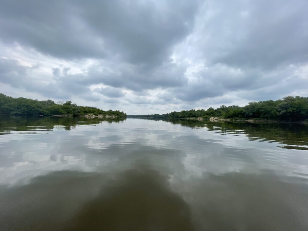 Szeroka, spokojna rzeka rozciąga się w oddali pod zachmurzonym niebem. Zielone drzewa rosną wzdłuż obu brzegów, a chmury i wierzchołki drzew wyraźnie odbijają się na gładkiej powierzchni wody, tworząc spokojny i symetryczny krajobraz.
