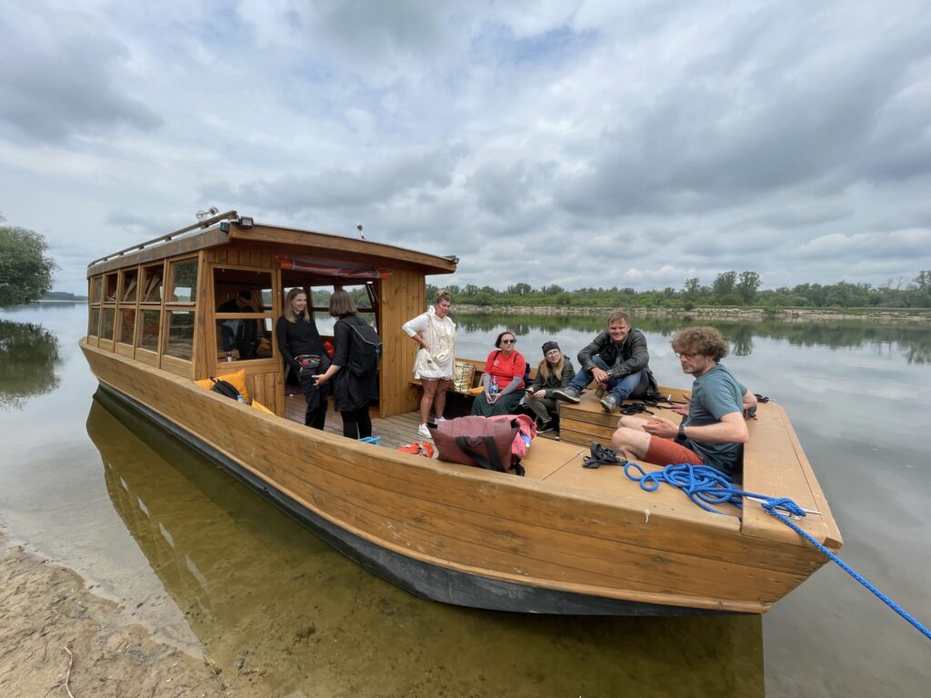 Grupa ludzi, niektórzy siedzący, a niektórzy stojący, gromadzą się na drewnianej łodzi zadokowanej na brzegu spokojnej, czystej rzeki. Niebo jest zachmurzone, a w tle widoczny jest brzeg rzeki z drzewami. Atmosfera wydaje się zrelaksowana i swobodna.