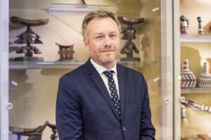 Mężczyzna z jasnobrązowymi włosami i krótką brodą, ubrany w granatowy garnitur, białą koszulę i wzorzysty krawat, stoi przed szklanymi półkami prezentującymi różne tradycyjne artefakty i rzeźby w otoczeniu przypominającym muzeum.