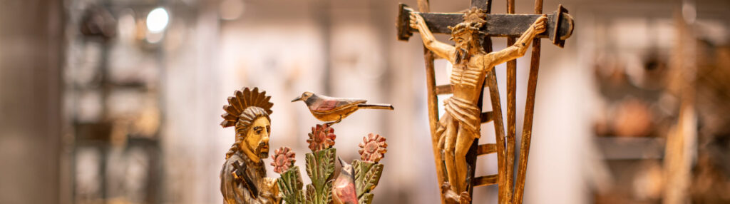 Zbliżenie drewnianej rzeźby ludowej przedstawiającej Jezusa na krzyżu z drabiną za nim. Po lewej stronie postać z pierzastym nakryciem głowy stoi wśród rzeźbionych kwiatów i ptaka. Tło jest delikatnie rozmyte, podkreślając szczegółowe rzeźby.