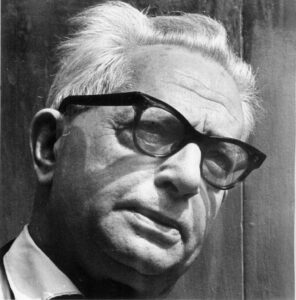 Czarno-białe zdjęcie starszego mężczyzny z jasnymi włosami, noszącego grube okulary w ciemnych oprawkach. Ma poważny wyraz twarzy i jest ubrany w marynarkę i koszulę z kołnierzykiem. Tło jest gładkie i zacienione.